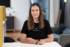 Porträt einer Frau mit langen braunen Haaren, die an einem Tisch sitzt. Sie trägt ein schwarzes T-Shirt mit dem Logo "FRIENDVENTURE" und einen Armband aus Gold und Blau. Im Hintergrund ist ein moderner Büroraum zu sehen.