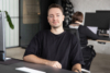Portrait des SEO Consultants Alex Kiritschenko, der in einem modernen Büro an einem Schreibtisch sitzt. Er trägt ein schwarzes T-Shirt und lächelt freundlich in die Kamera.