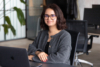 Portrait einer Frau mit schulterlangen, welligen braunen Haaren, die in einem modernen Büro an einem Schreibtisch sitzt. Sie trägt einen grauen Strick Cardigan und eine Brille. Ihre Arme sind vor ihr auf dem Tisch verschränkt. Im Hintergrund sind Pflanzen und Schreibtische zu sehen. Die Büroatmosphäre ist minimalistisch und modern.