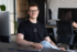 Ein junger Mann mit kurzem braunen Haar und schwarzem T-Shirt sitzt entspannt an einem Schreibtisch in einem modernen Büro. Er blickt freundlich in die Kamera. Im Hintergrund arbeitet eine weitere Person an einem Laptop, umgeben von Monitoren und Büromöbeln.