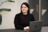 Porträt einer Frau mit langen dunklen Haaren und goldenen Creolen, die an einem Schreibtisch vor einem geöffneten MacBook sitzt und in die Kamera lächelt. Im Hintergrund sind eine grüne Zimmerpflanze und unscharfe Büroelemente zu sehen.
