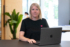 Porträt einer Frau mit blonden Haaren in einem schwarzen T-Shirt. Sie sitzt an einem Tisch in einem modernen Büro mit Pflanzen im Hintergrund und lächelt in die Kamera.