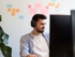 Ein junger Mann mit Kopfhörern arbeitet konzentriert an einem Computerbildschirm. Im Hintergrund sind bunte Post-its an der Wand zu sehen, daneben eine grüne Pflanze.