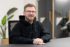 Michael, unser Developer, sitzt in an einem Bürotisch, er trägt einen schwarzen Hoodie und eine Brille. Seine Hände liegen auf dem Tisch und er lächelt freundlich in die Kamera. Im Hintergrund sieht man Call-Kabinen und Pflanzen.