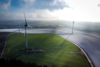Wind turbines standing on green and snow-covered fields under a cloudy sky.