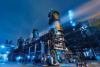 Industrial plant with metal structures and pipelines illuminated at night under a blue sky.