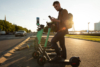 Man standing on an e-scooter at sunset, checking his phone near a row of parked scooters.