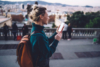Woman with a backpack smiling while using her smartphone outdoors with a city view in the background.