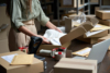 Person packing a cardboard box for shipping in a warehouse, surrounded by parcels and packing materials.