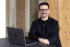 Chris, unser TYPO3 Lead sitzt in unserem Office an einem Tisch, vor ihm steht ein Macbook und er lacht freundlich in die Kamera. Er hat dunkle Haare, trägt eine Brille und ein dunkles zugeknöpftes Hemd.