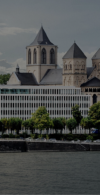 Eine Panoramaaufnahme der Kölner Skyline am Rheinufer bei bewölktem Himmel. Im Zentrum steht ein modernes, weißes Bürogebäude mit einer gleichmäßigen Fassadenstruktur direkt hinter der Uferpromenade. Rechts daneben ragen die markanten Türme der romanischen Kirche Groß St. Martin auf. Am unteren Bildrand ist ein weißes Passagierschiff auf dem Rhein zu sehen.